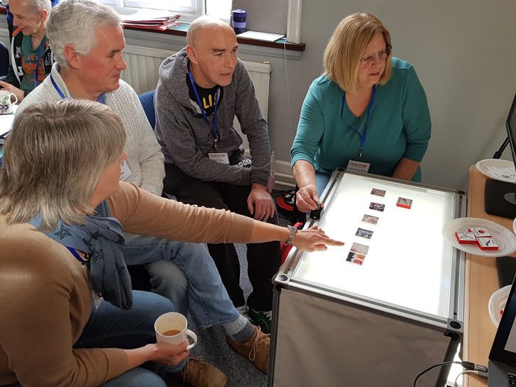 Four adults are seated around a white table frame which is lit from beneath to make the table surface glow white.  There are a series of small square pictures on the tabletop. All adults are looking towards a computer monitor just out of shot. One participant is adjusting a small dial at the front of the tabletop and another is pointing towards one of the small pictures arranged in a row on the table.