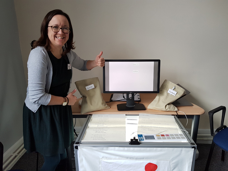 Abi, a white female with long brown hair, stands next to a white table frame which lit from beneath to make the table surface glow white. Behind the table frame sits a computer monitor on a side table. Abi is smiling at the camera and giving a thumbs up gesture.